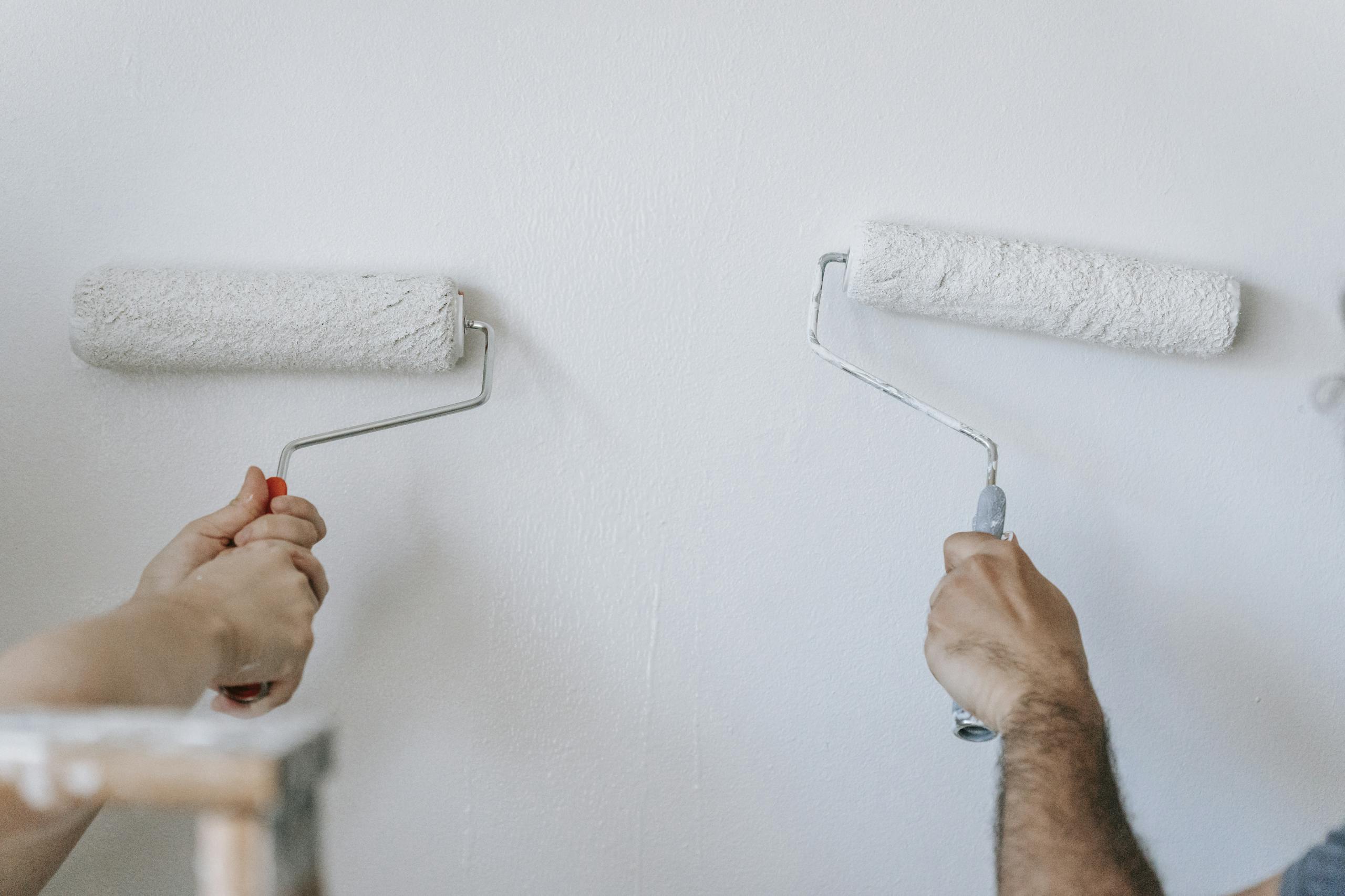 Close-up of two individuals painting a wall with paint rollers indoors.